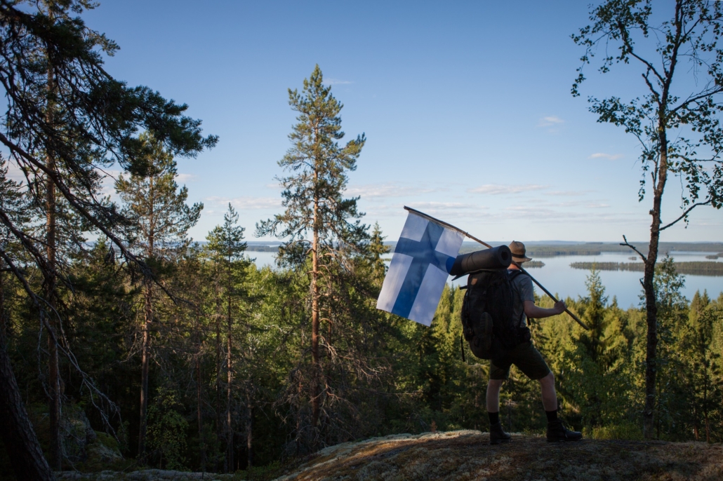 Retkeilijä katselee järvimaisemaa korkealta kalliolta Suomen lippua kannatellen. Kuva Joel Heino/Metsähallitus