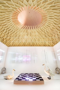 Taideteos puisen rakennelman sisällä, jossa kaksi joutsenta seisoo shakkipelin äärellä