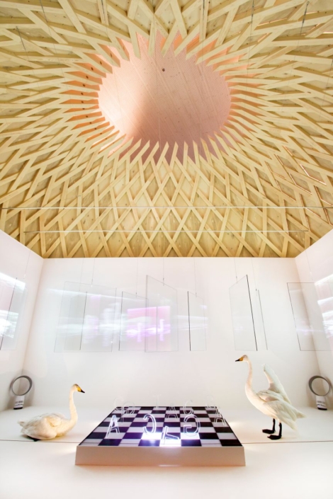 Taideteos puisen rakennelman sisällä, jossa kaksi joutsenta seisoo shakkipelin äärellä